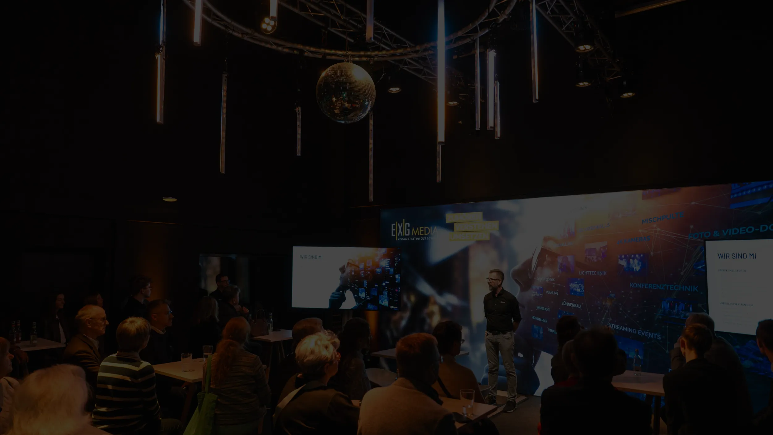 Das Bild zeigt eine Bühne mit einem Speaker, vor einem Backdrop stehend, links und rechts davon ein Display. Stehend vor Zuschauern